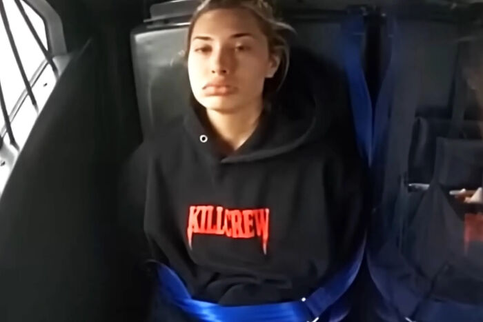 Young woman wearing a black hoodie seated in a vehicle, husband stands by side of teacher accused of sexual acts at school.