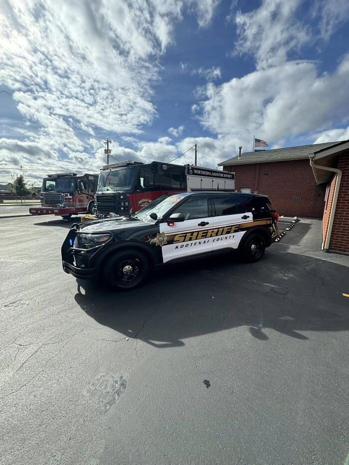 Sheriff vehicle parked near fire trucks outside a Northern Lakes Fire District building under a partly cloudy sky.