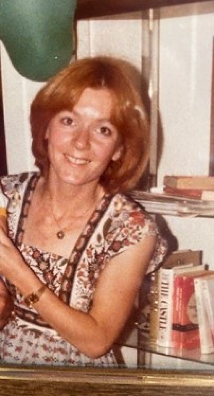 Smiling woman with red hair and floral dress in a home setting with books, related to cold case murder of school teacher.