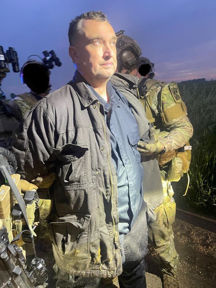 Alleged shooter from 2025 crimes escorted by armed military personnel outdoors at dusk in a dark jacket.