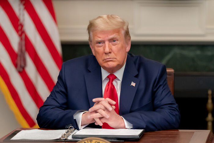 Donald Trump sitting at desk with folded hands, focused expression, U.S. flag in background, related to probe of Biden autopen use.