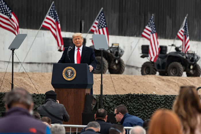 Former President Trump speaking at a military parade with American flags and armed forces vehicles in the background.