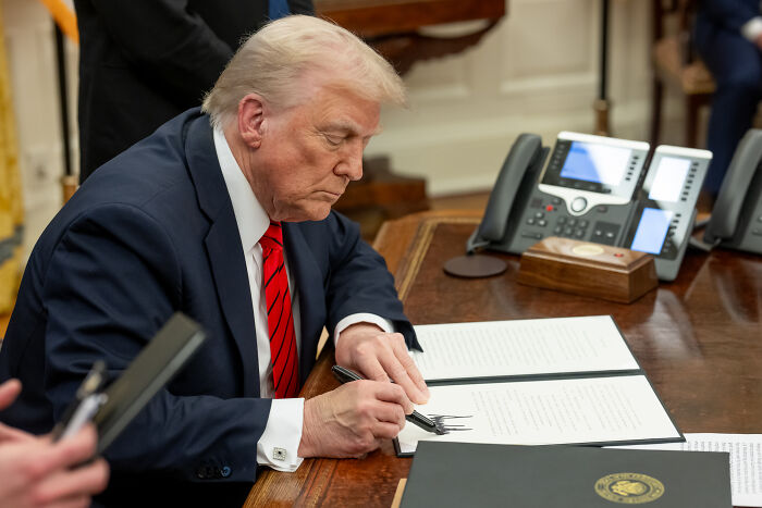 Former President Trump in a navy suit and red tie signing documents at a desk, related to Jan 6 rioter pardon proposal.