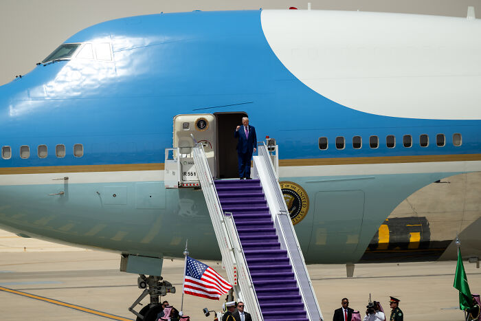 Donald Trump descending Air Force One steps, stumbling and dressed in a dark suit with an American flag nearby on the tarmac.