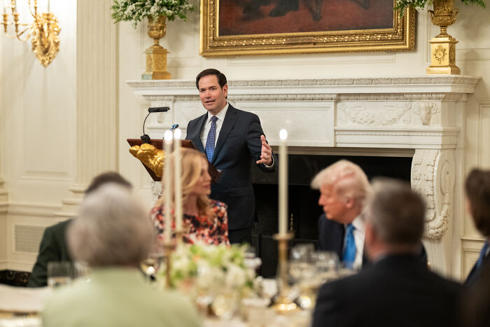 Man in dark suit speaking at a formal dinner, with attendees listening, referencing Elon Musk and foreign student ban topics.