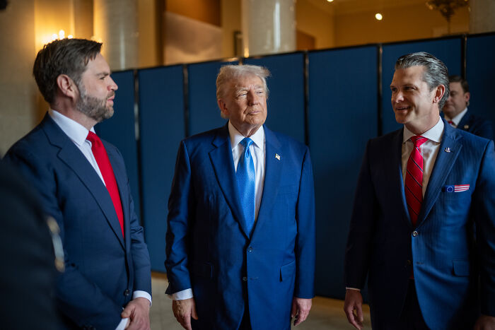 Donald Trump in a blue suit signing a travel ban limiting entry from 19 countries, standing with two men in suits.