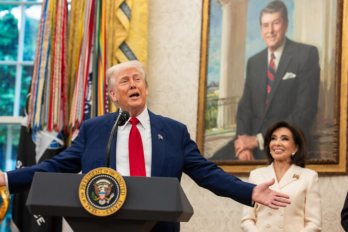 Former US president speaking at a podium with presidential seal, with a woman smiling and a portrait in the background.