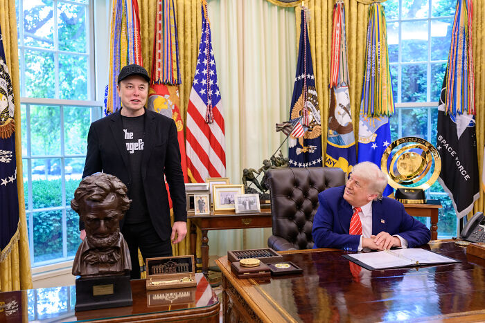 Elon Musk standing beside Donald Trump in an office setting with American flags and presidential decor in the background.