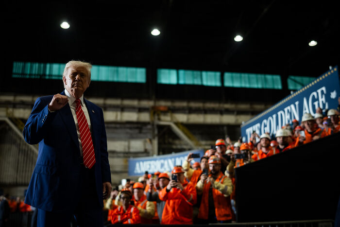 Man in a suit standing in front of a crowd wearing orange safety gear during an indoor industrial event.