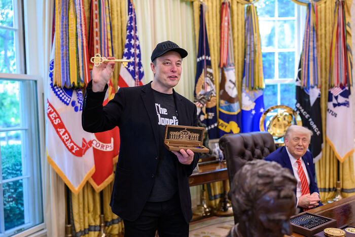 Elon Musk holding a key and model in Oval Office, surrounded by military flags, amid foreign student ban discussions.