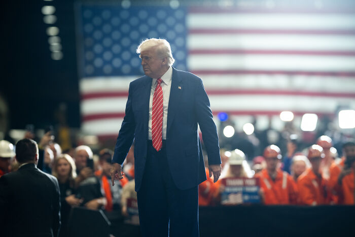 Donald Trump at a public event with a large American flag, related to travel ban limiting people from 19 countries.