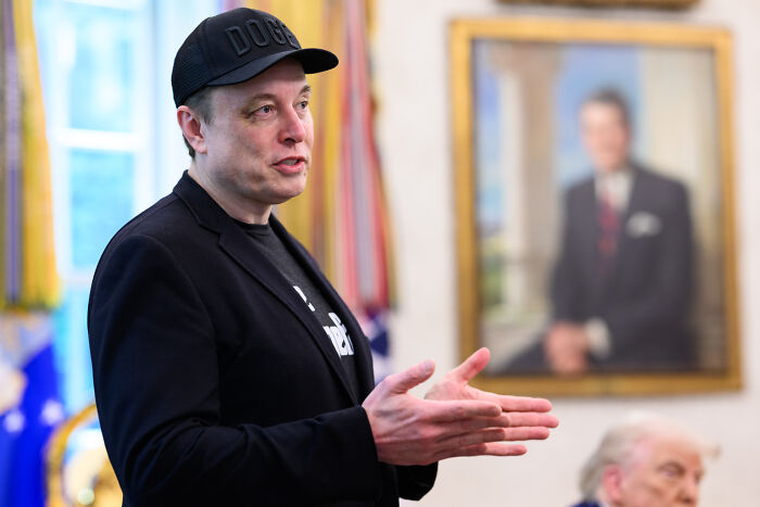 Elon Musk speaking indoors wearing a black cap and jacket, with a blurred portrait and Donald Trump in the background.