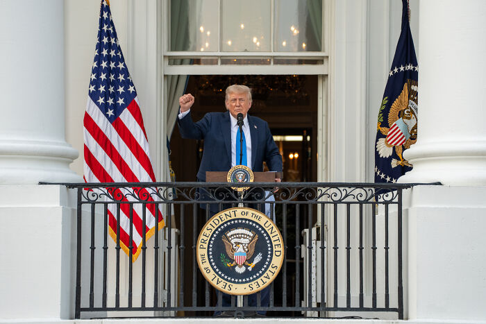 Former President Trump speaking from a White House balcony with US flags during a political event.