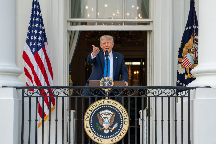 Donald Trump speaking on White House balcony with presidential seal and American flags during a public address.