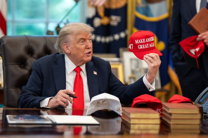 Donald Trump sitting at desk in Oval Office holding a red Make America Great Again hat, related to Air Force One steps stumble.