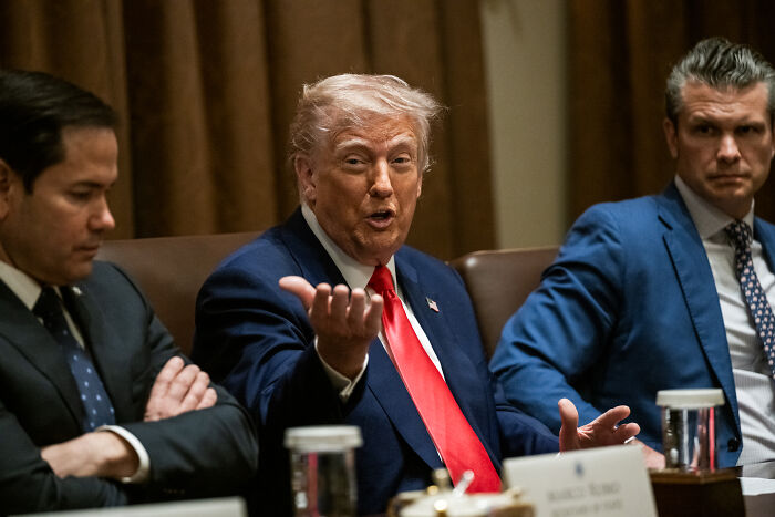 Donald Trump speaking at a meeting, gesturing with hand, related to military crackdown and political tensions in Los Angeles.