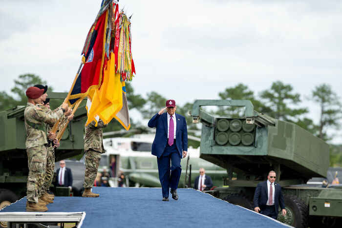 Man in a suit saluting near soldiers holding flags at a military event contrasting low-energy birthday parade and no kings rally.