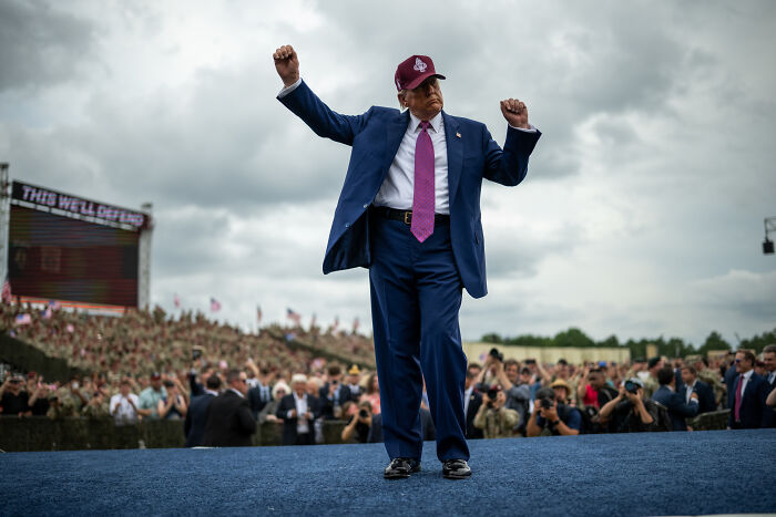 Donald Trump in a blue suit and red hat raising fists at a public event amid a large crowd in an outdoor setting.