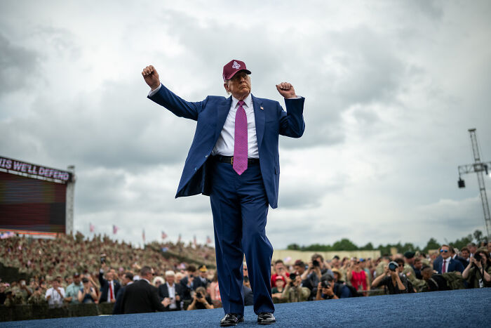 Donald Trump reacts at a public event wearing a suit and cap, engaging with a large crowd under a cloudy sky.