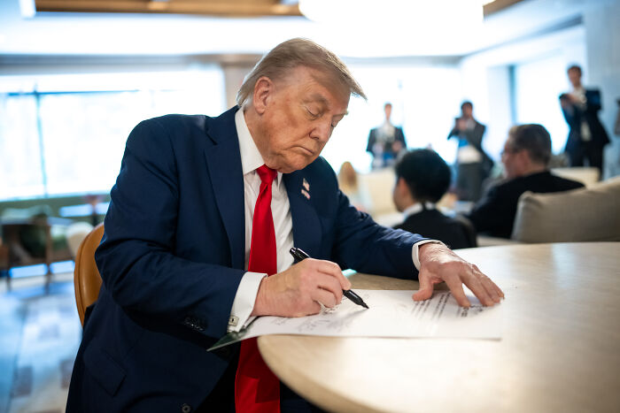 Donald Trump in a dark suit and red tie signing documents at a table amid a formal gathering, related to Iran attack plan approval.