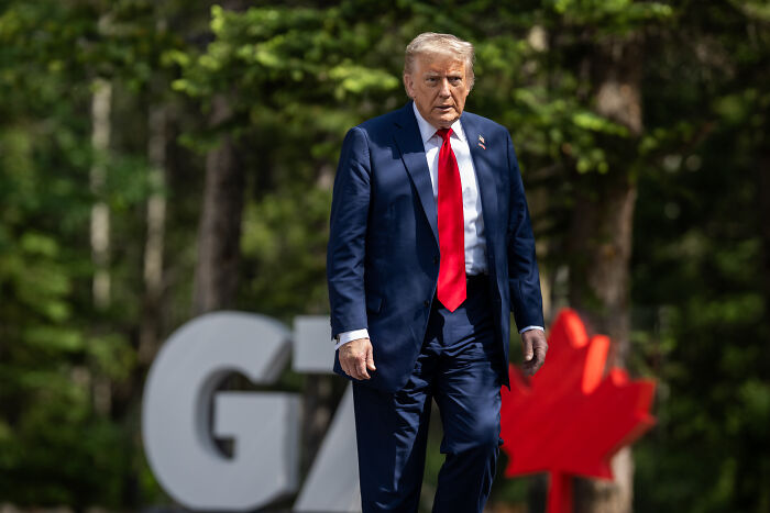 Man in a navy suit and red tie outdoors near a G7 sign, symbolizing Vice President JD Vance suspended from Bluesky platform.