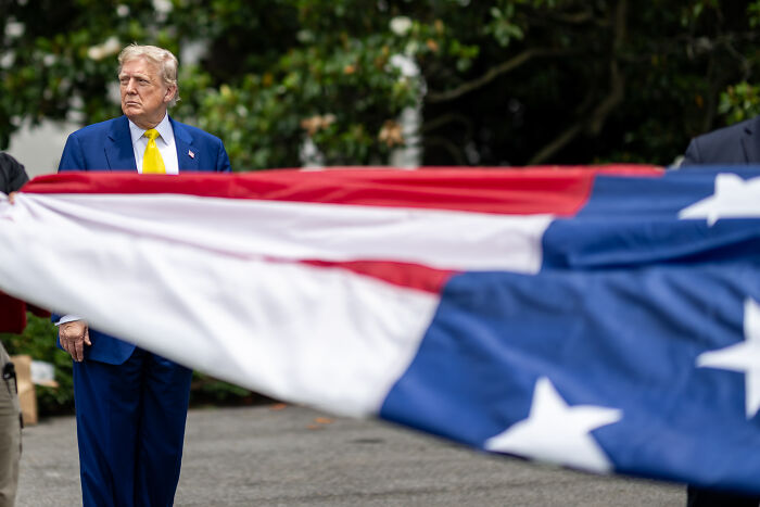 Former President Trump in a blue suit standing near a large American flag amid discussions on White House Juneteenth acknowledgment.