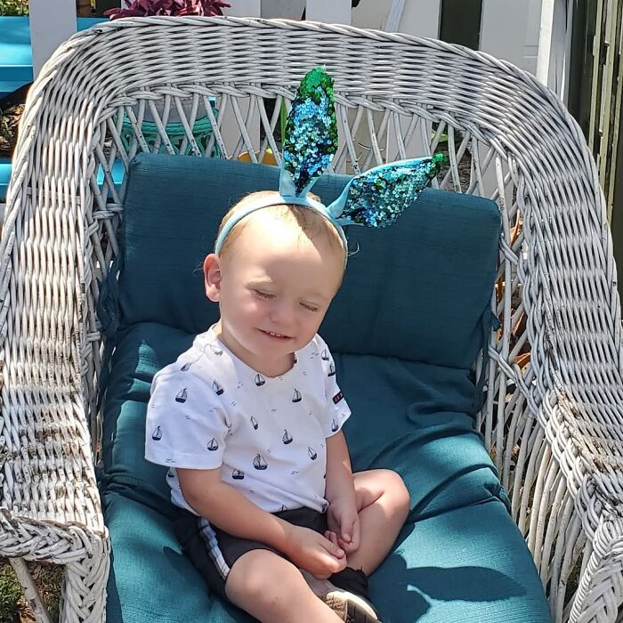 Baby boy wearing blue sequin bunny ears sitting on a cushioned wicker chair outdoors in Florida.
