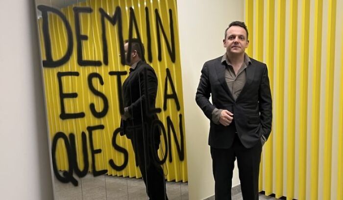 Man in black suit standing beside a mirror reflecting graffiti, related to Sarco Pod advocate and capsule incident.