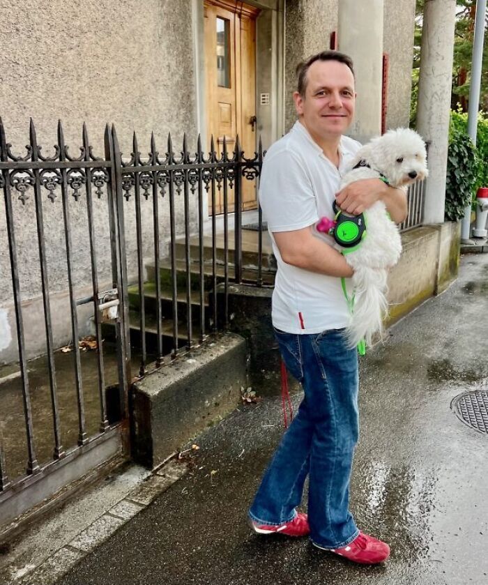 Man holding white dog outside near metal fence, related to Sarco Pod advocate and capsule death news.