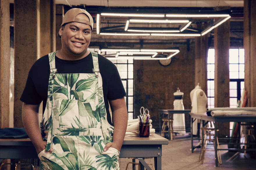 Fashion designer standing in a studio wearing tropical print overalls, smiling with sewing tools and mannequins in background.