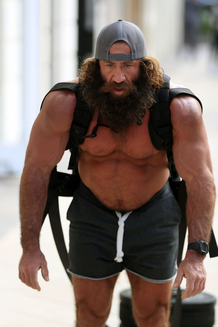 Muscular man known as Liver King wearing a backward cap and carrying heavy weights during outdoor workout session.