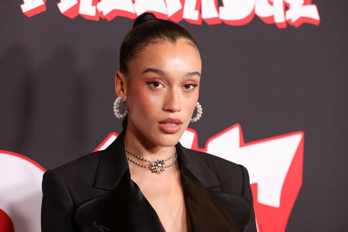 Singer wearing earrings and black blazer at event, linked to Dominican jersey and singing US anthem in Spanish controversy.