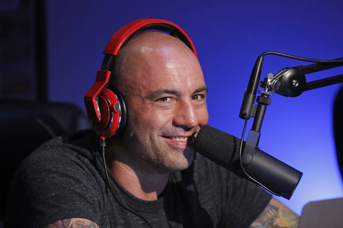Podcaster Joe Rogan smiling with red headphones speaking into a microphone in a dimly lit studio.