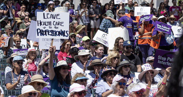 Crowd at a protest holding signs about healthcare professionals and political interference in emergency care under Florida abortion law.