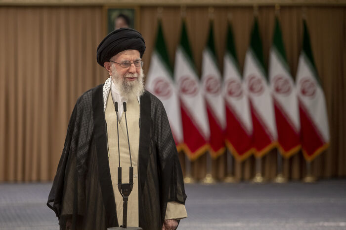 Iran&rsquo;s Supreme Leader speaking at a podium with multiple Iranian flags in the background during a public statement.