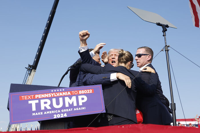Trump assassination attempt aftermath with security and staff in protective embrace during public event outdoors.