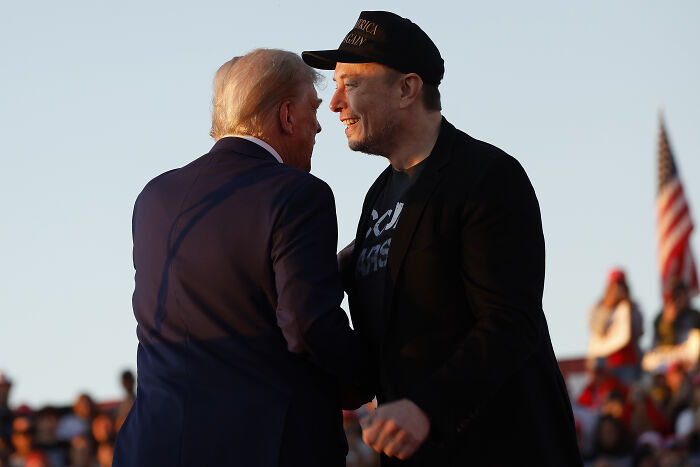 Elon Musk and Donald Trump shaking hands outdoors at an event with American flags in the background.