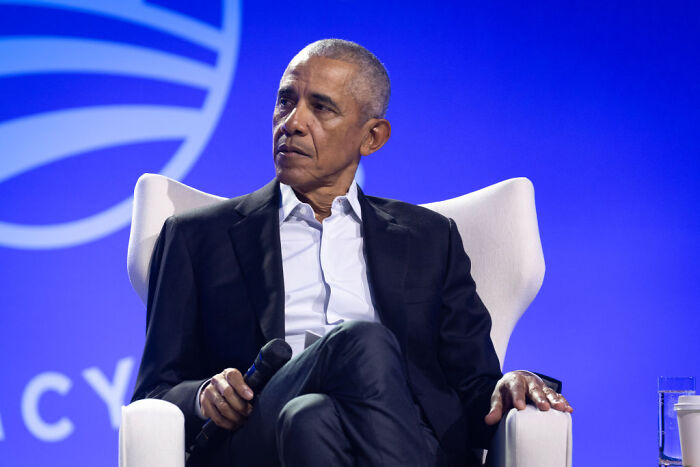 Barack Obama seated on stage holding a microphone, discussing topics related to MAGA hypocrisy in a formal event setting.