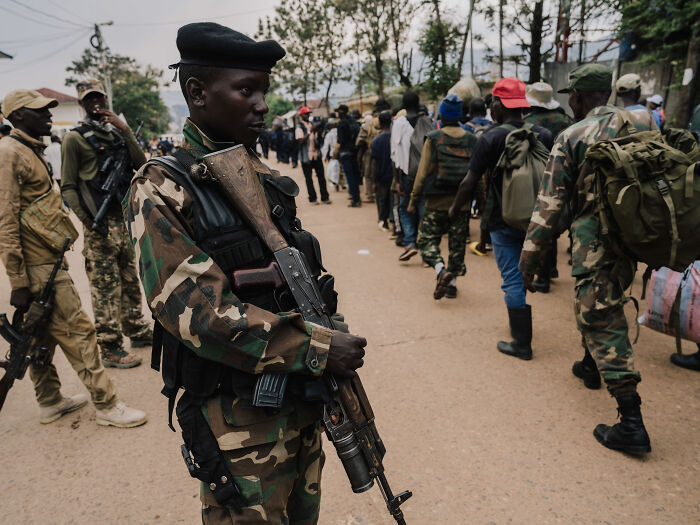 Armed soldier in camouflage standing guard as civilians and soldiers walk, highlighting deadly wars and humanitarian crises.