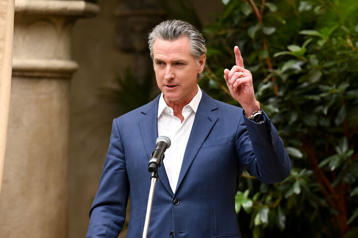 California Governor speaking at a podium outdoors with greenery and stone columns in the background about National Guard control.