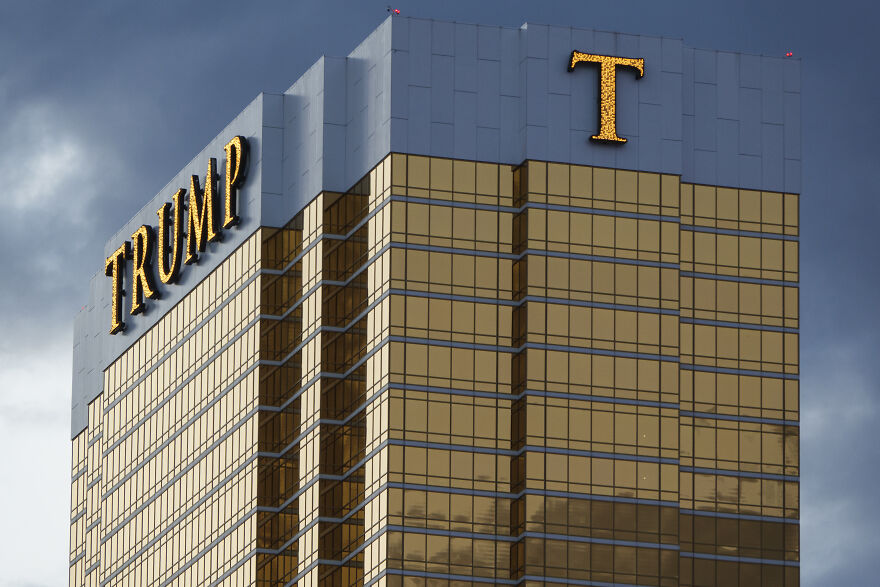 Trump Organization building with gold reflective windows and illuminated Trump logo against a cloudy sky background.