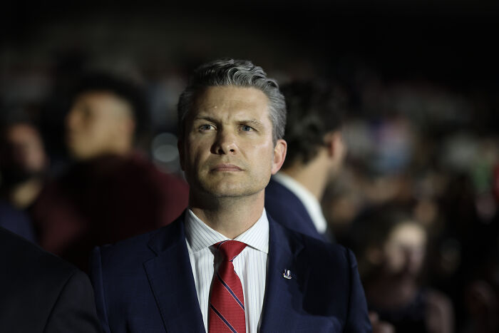 Pete Hegseth wearing suit and red tie, attending event while Navy vessel rename debate involving Harvey Milk unfolds.
