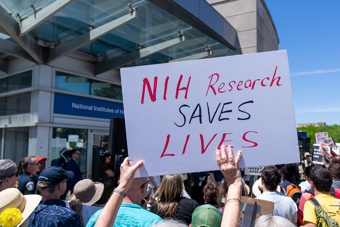 Crowd protests outside National Institutes of Health building with sign supporting NIH research and grant reinstatement.