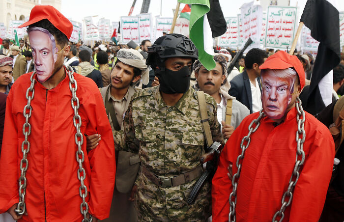 Protester in a war zone wearing political masks and orange jumpsuits with chains, highlighting deadly wars and humanitarian crises.