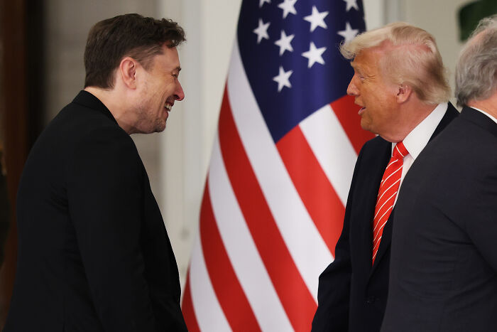 Elon Musk and Donald Trump laughing and talking in front of an American flag during a public event.
