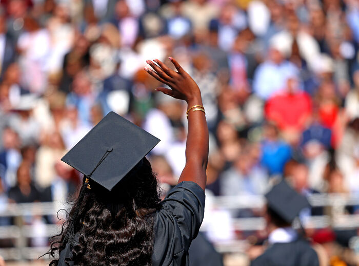 Graduate wearing cap and gown raising hand at ceremony with crowd in the background, representing foreign student visas.