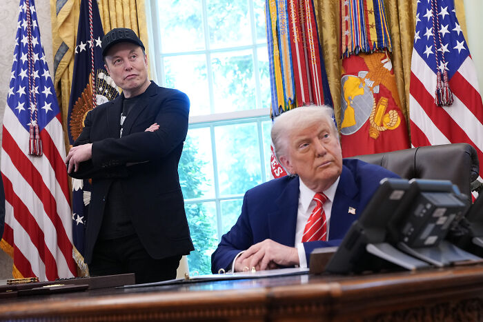 Elon Musk standing with arms crossed beside Donald Trump seated at desk in an office with flags and decorations.