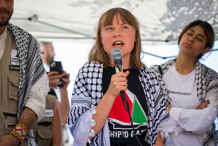 Greta Thunberg speaking into a microphone aboard an aid ship, surrounded by people wearing keffiyeh scarves.