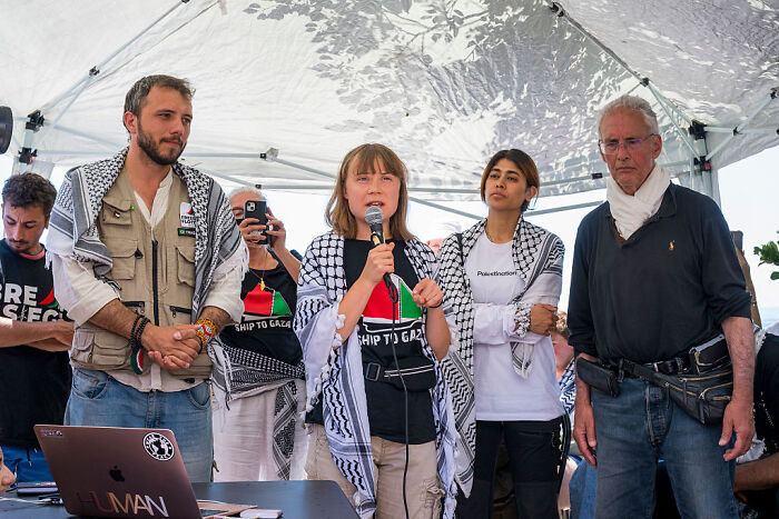 Greta Thunberg speaking during a press event aboard aid ship, surrounded by activists wearing Palestinian scarves.