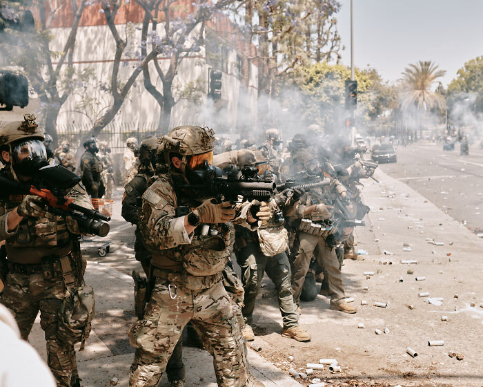 Armed military personnel in tactical gear advancing on a smoke-filled street amid LA riots and unrest.